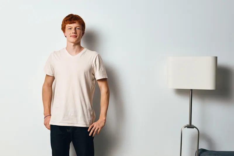 Young man with red hair stands against plain white wall indoors wearing white t shirt and dark jeans with relaxed confident pose and soft lighting in modern room interior.