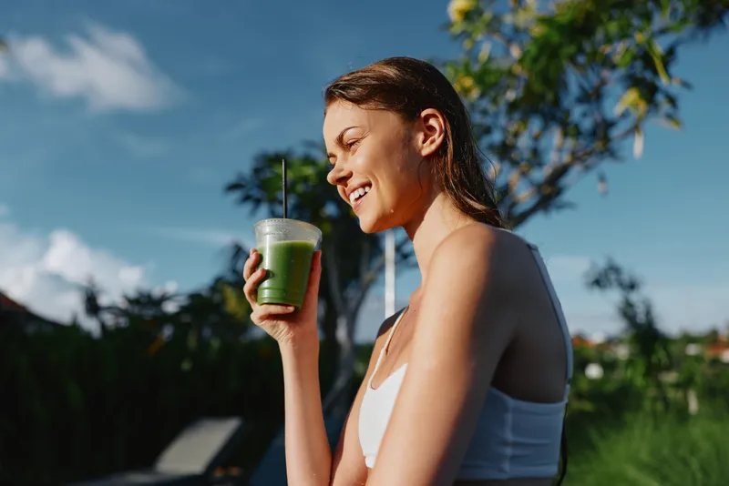 A young woman in a white top drinks a green smoothie outdoors, enjoying sunshine and fresh air. A lifestyle moment focused on wellness, hydration, and daytime outdoor vitality.