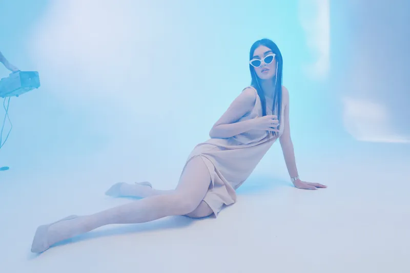 Fashionable woman in a beige dress and sunglasses poses on the floor in a bright studio with soft blue lighting. Concept for beauty, fashion, and modern style photography.