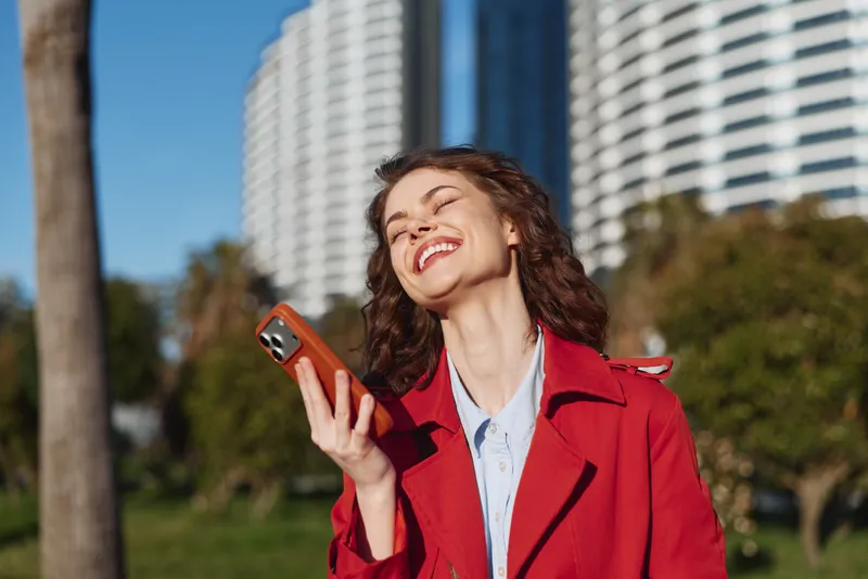 Young woman with curly hair wearing a red coat smiles joyfully while holding a smartphone outdoors on a sunny day near modern buildings and green trees.