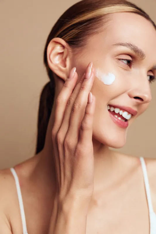 Young woman with clear skin applies moisturizer cream on face smiling gently isolated on beige background in skincare beauty routine concept for healthy glowing skin care.