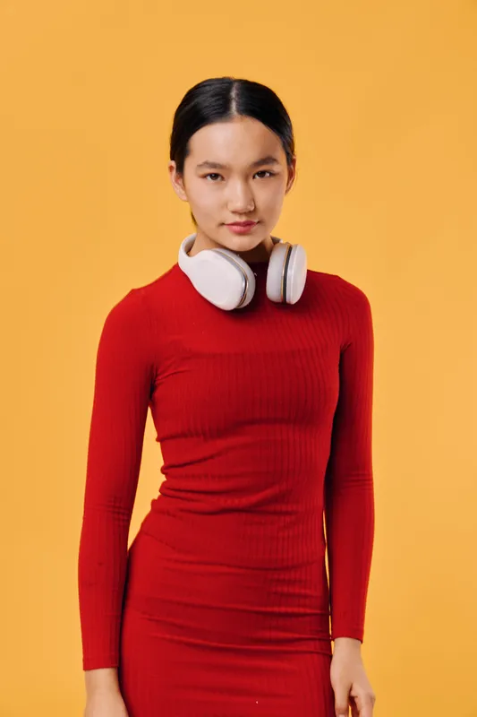 Portrait of young woman in red dress with white headphones around neck standing against orange background looking at camera with calm expression and straight posture.
