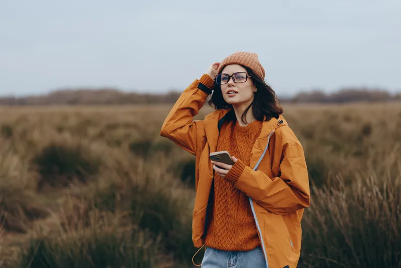 Woman lifestyle outdoors field, wearing hat and glasses, orange jacket and knit sweater, holding phone, relaxed pose, warm autumn light, blurred grass background, soft breeze natural colors