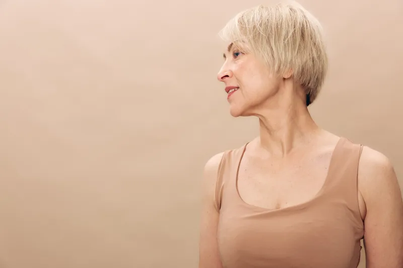 Portrait of a mature woman with short blonde hair wearing a sleeveless top against a beige background. Concept of aging, natural beauty, and thoughtful expression.