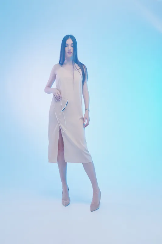Young woman in beige dress stands confidently holding earphones in a minimal studio with soft blue lighting. Concept of woman beauty fashion and modern lifestyle.