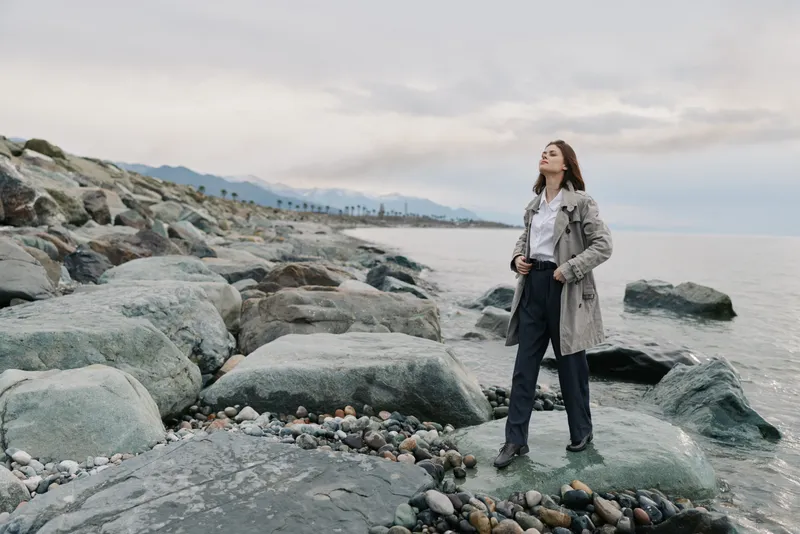 Young woman stands confidently on rocky shore wearing casual coat and jeans. Cloudy sky and calm sea create peaceful atmosphere. Concept of nature, freedom, and outdoor lifestyle.