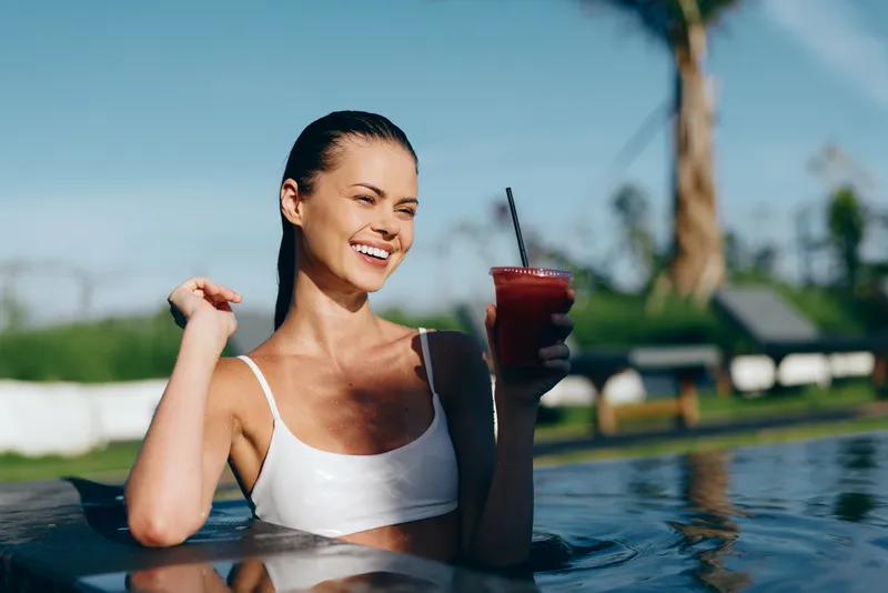 Young woman in a white swimsuit enjoys a refreshing drink while relaxing in a swimming pool on a sunny day with a joyful and carefree expression.