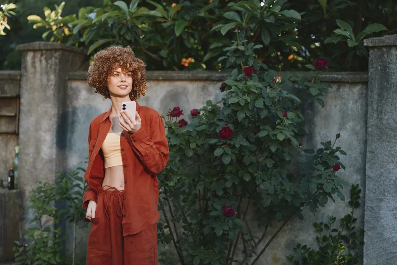 Curly haired woman wearing a rust colored suit poses with a smartphone in a sunny garden, capturing a casual lifestyle moment, outdoor fashion, and personal expression. The scene emphasizes natural