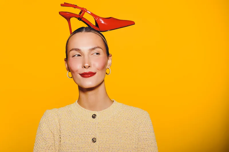 Young woman with red high heel shoes on her head smiles and looks sideways. Bright yellow background and stylish beige outfit create playful and creative fashion concept.