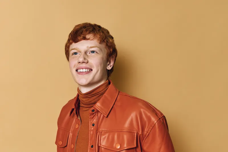 Portrait of smiling young man with red hair wearing orange leather jacket and turtleneck standing against beige background, happy expression and casual style in studio light.