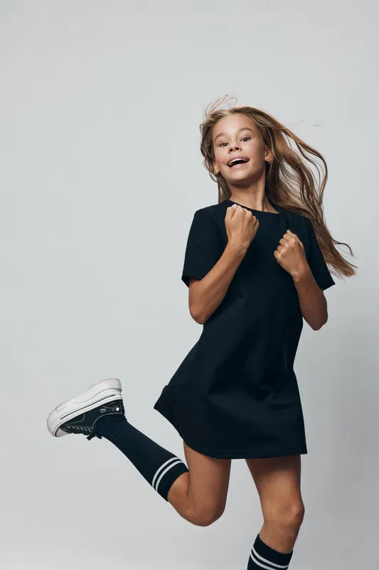 Girl jumping and smiling with black dress and sneakers on white background. Child with long hair, happy emotion, casual outfit and active mood in studio.