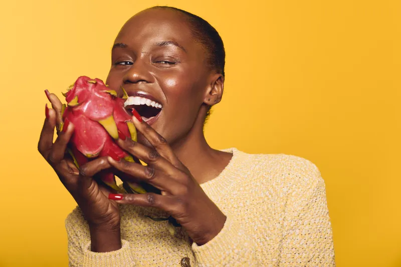 Smiling dark skinned African woman in beige sweater holds ripe pink dragon fruit near mouth on yellow background. Concept of healthy eating, exotic fruit, natural lifestyle and nutrition.