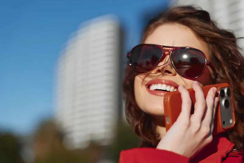 Young woman with curly hair wearing sunglasses smiles while talking on a smartphone outdoors on a sunny day in the city.