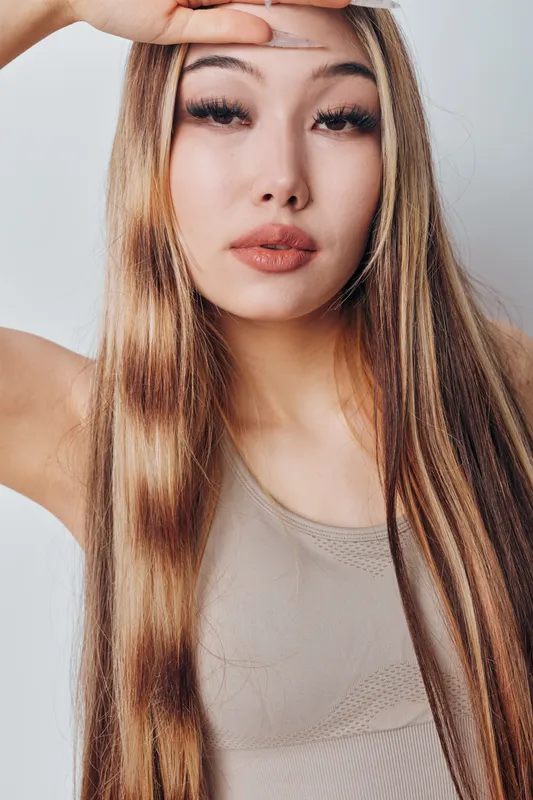 Woman with long hair and natural makeup looking at camera posing with hand on forehead in studio portrait against white background with soft lighting.