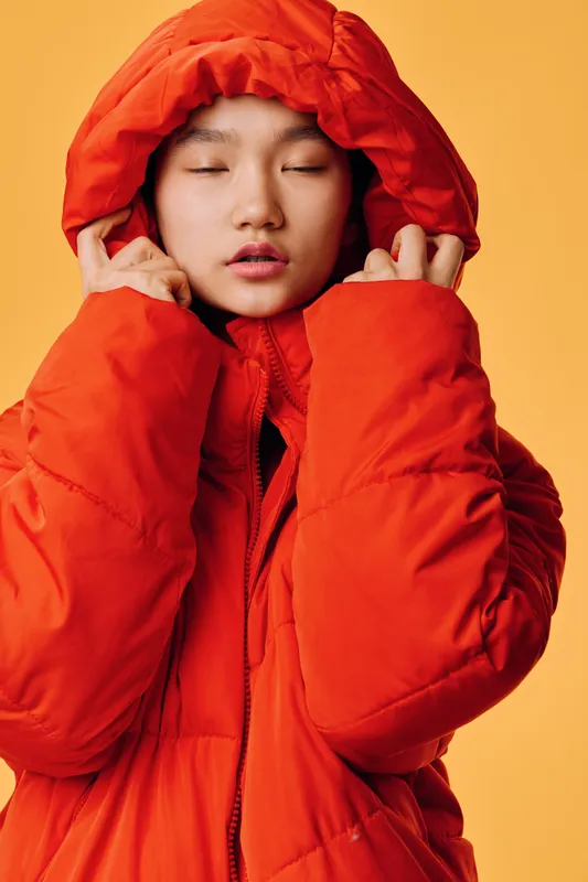 Woman in orange puffer jacket with hood up, eyes closed, posing against yellow background. Fashion, winter clothing, casual style, warm wear, cold weather outfit, beauty and comfort concept.