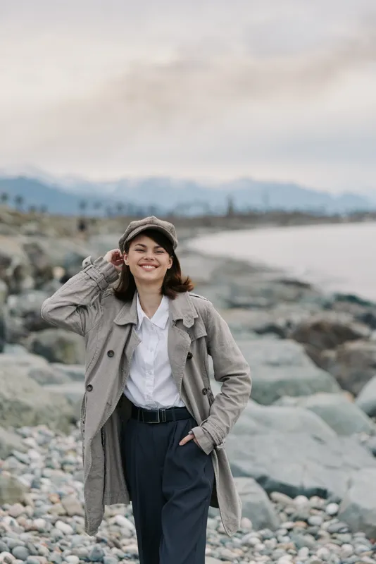 Smiling woman in knitted coat and cap walks on rocky beach near sea under cloudy sky. Autumn fashion concept with casual outfit and natural outdoor lifestyle by the coast.