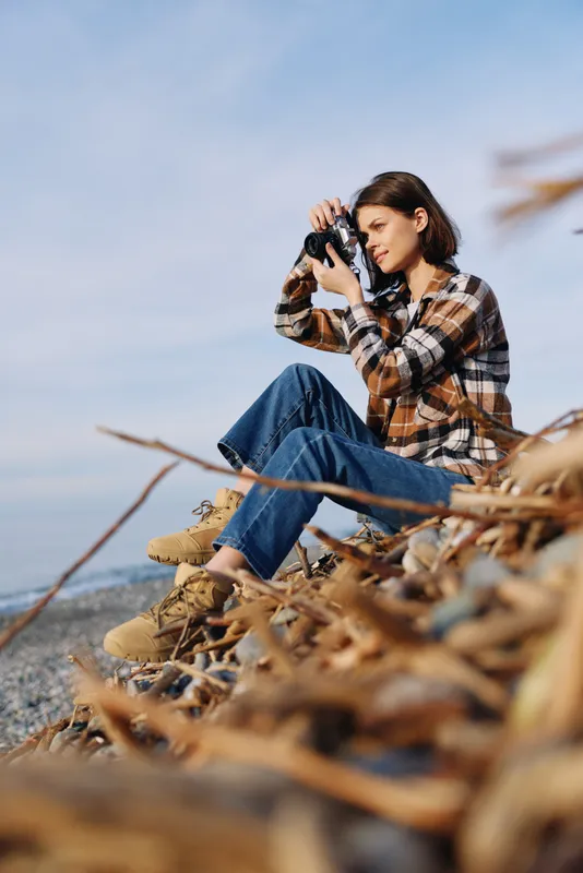 Woman photographer sitting on the beach among dried grasses, using a camera to capture the seascape under bright daylight, wearing a plaid shirt and jeans.