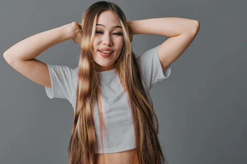 Young woman with long brown hair wearing white crop top standing with hands behind head and smiling with closed eyes on gray background.