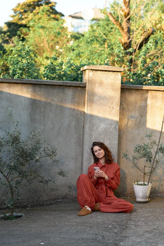 Relaxed woman wearing a rust red outfit sits on a concrete patio beside a tall wall, browsing a smartphone. Warm daylight and soft shadows create a casual lifestyle mood with natural outdoor ambiance
