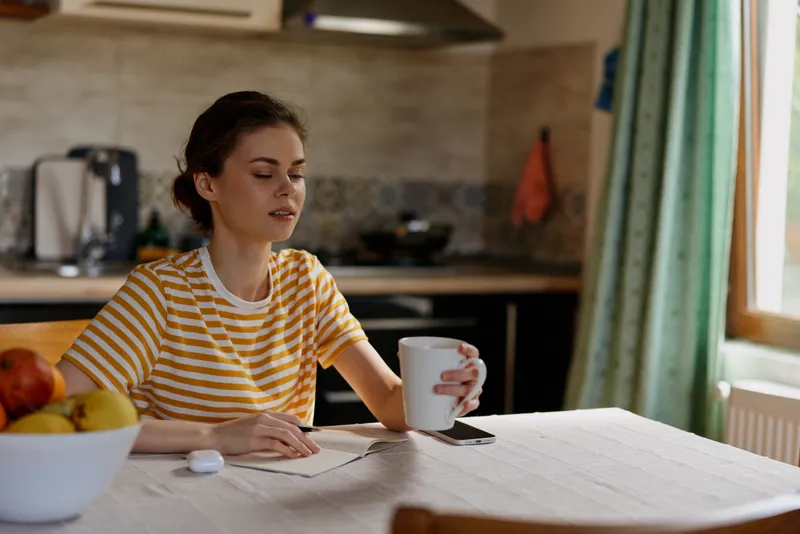 Candid woman lifestyle at home, seated at a bright kitchen table, writing in a notebook, holding a mug, daylight streaming in, calm, organized morning routine.