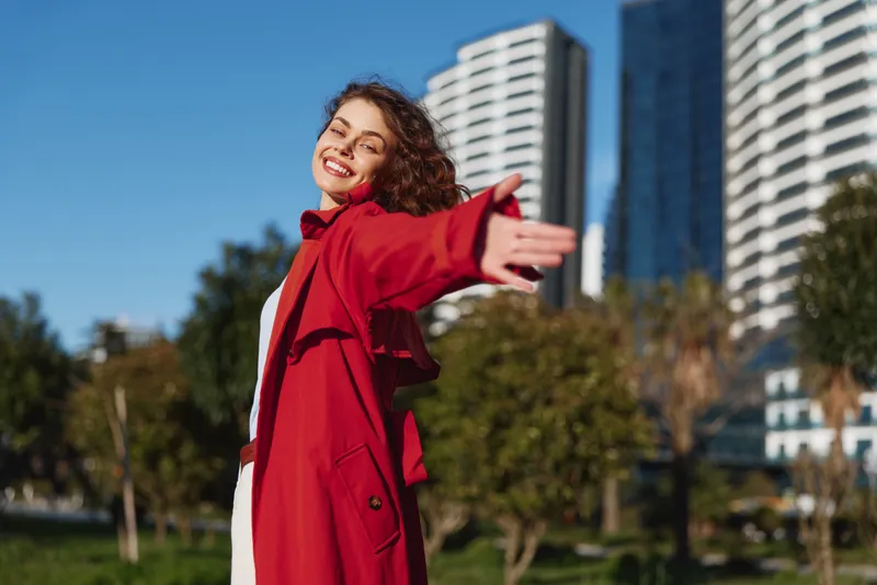 A woman in a red coat stands in a city park with open arms, exuding warmth and confidence as modern buildings rise behind her, a moment of bold self-expression in bright daylight.
