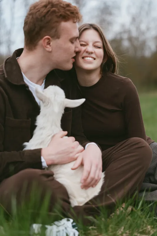 Couple outdoors with a goat kid, smiling woman and man kissing her cheek in a meadow. Casual lifestyle moment, warm natural lighting, affectionate bond, playful interaction with animal, relaxed,