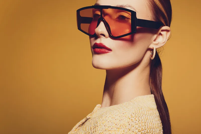 Stylish woman with long hair wearing oversized orange sunglasses and red lipstick poses against yellow background. Fashion portrait with modern accessories and confident expression.