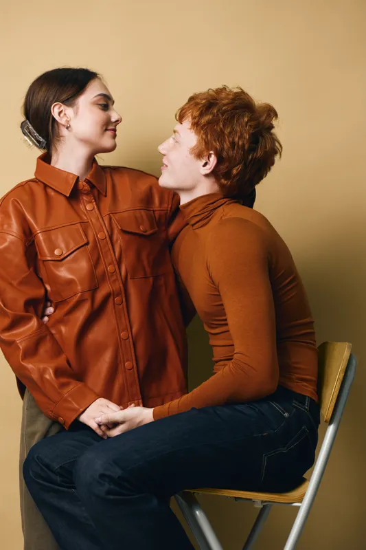 Couple, love, and relationship with man sitting on chair holding woman hands, looking at each other. Intimate moment with affection and connection indoors against beige background.