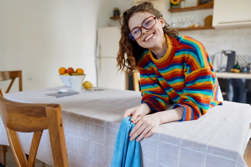 Cheerful woman indoors, smiling, colorful striped sweater, relaxed pose at dining table, sunlit kitchen vibe, casual lifestyle, warm hues, home comfort, candid moment