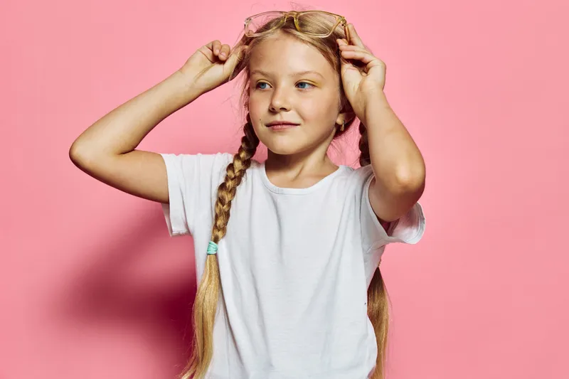 Young girl with pigtails wearing a white t-shirt adjusts her sunglasses on a pink background. Studio portrait with playful and confident mood.