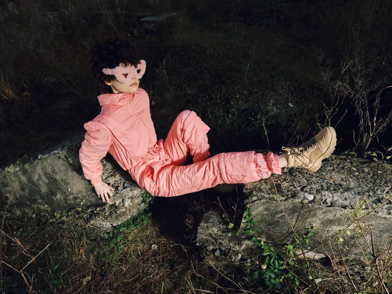 Mannequin in pink jumpsuit rests on rocky ledge at night, outdoors, boots on stones, dark foliage behind, eerie stillness and solitary figure in rugged landscape under pale moonlight and silent air.
