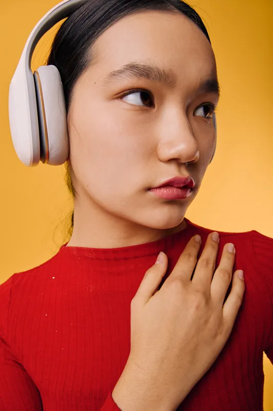 Young woman in red sweater wearing headphones standing against yellow background looking sideways with calm expression and hand placed on chest in closeup portrait style.