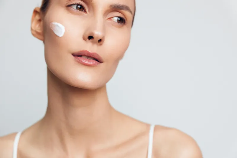 A woman with clear skin applies white face cream on her cheek while looking away. The closeup shot highlights skincare, beauty, and natural care concepts with soft lighting.