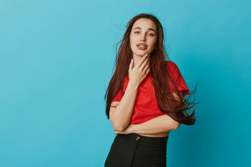 Young woman with long hair posing in red shirt and black pants on blue background. Casual style, confident expression, fashion portrait with natural makeup and relaxed posture in studio.