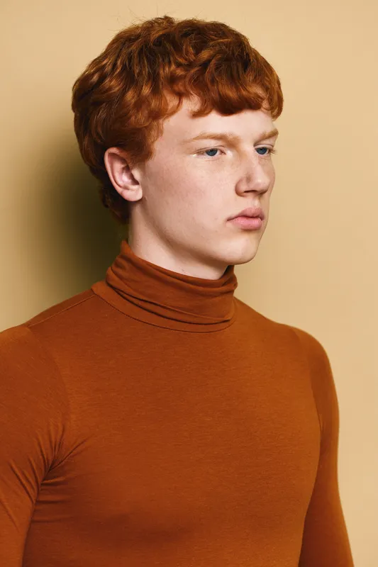Young man with red hair wearing a brown turtleneck looking sideways with neutral expression against beige background in a studio portrait photograph.