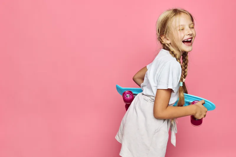 Happy young girl with long hair holding a blue skateboard laughing on pink background. Studio portrait of joyful child playing.