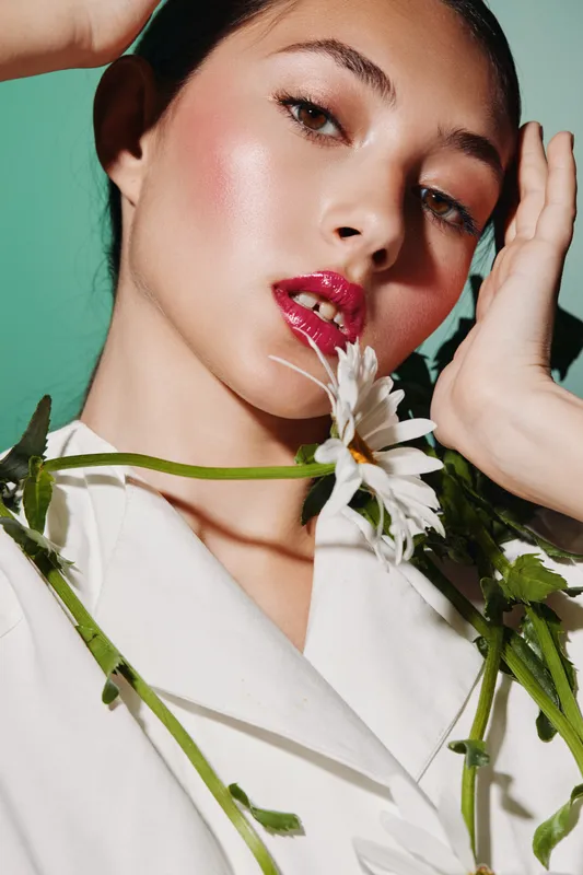 Portrait of a fashionable young woman with glossy lips holding white flowers near her chin against mint background. Concept for beauty, creative fashion, and floral aesthetics.