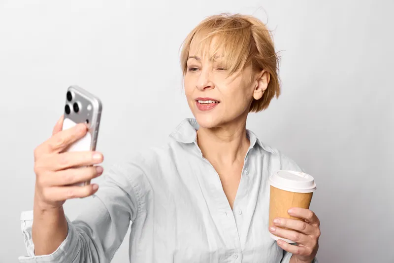 Woman with smartphone taking a selfie, smiling while holding a coffee cup, older portrait on bright white background, casual home lifestyle, soft studio light, today