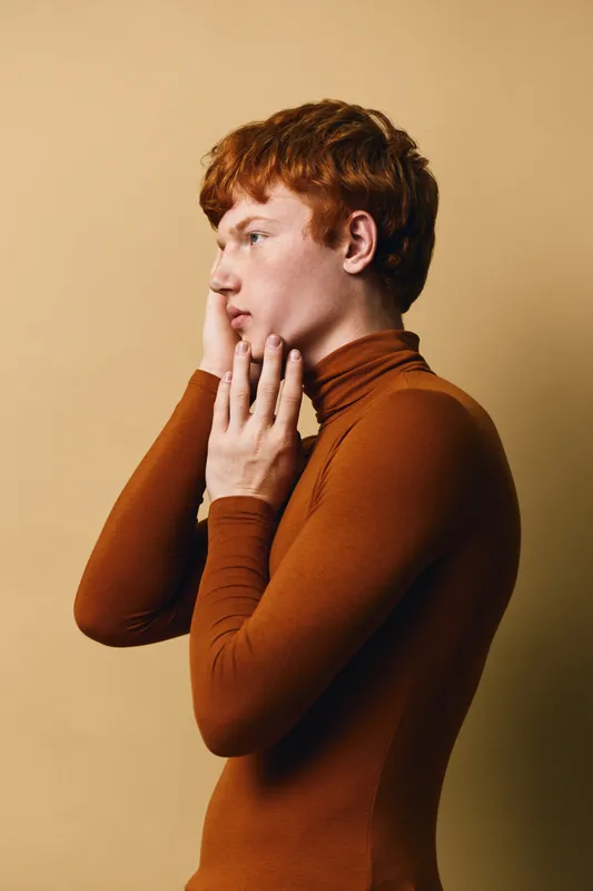 Young man with red hair and fair skin in brown turtleneck poses against beige background looking sideways with thoughtful expression and hand on face in profile view.