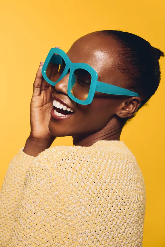Smiling African woman with dark skin wearing large blue sunglasses and a yellow sweater poses against a bright yellow background. Concept of fashion, joy, and summer style.