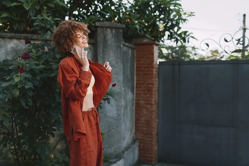Curly haired woman outdoors in a burnt orange suit adjusts her hair and gazes aside, a casual lifestyle portrait in a sunlit garden setting, warm and confident