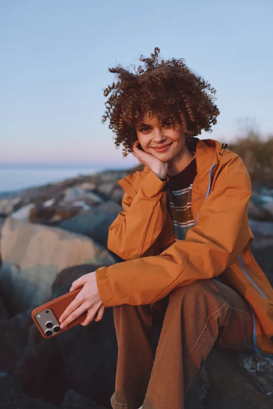 Woman lifestyle outdoors at seaside rocks during sunset breeze curly hair orange jacket smiling while holding phone for selfie view of ocean and sky shallow depth of field