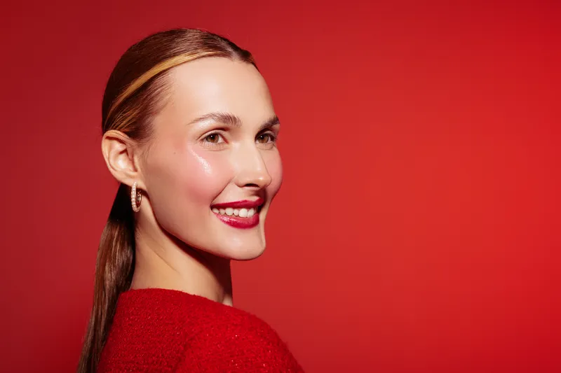 Smiling woman with red lipstick and sleek hair poses against red background. Studio portrait with bright lighting highlights her clear skin and confident expression. Concept of beauty and fashion.