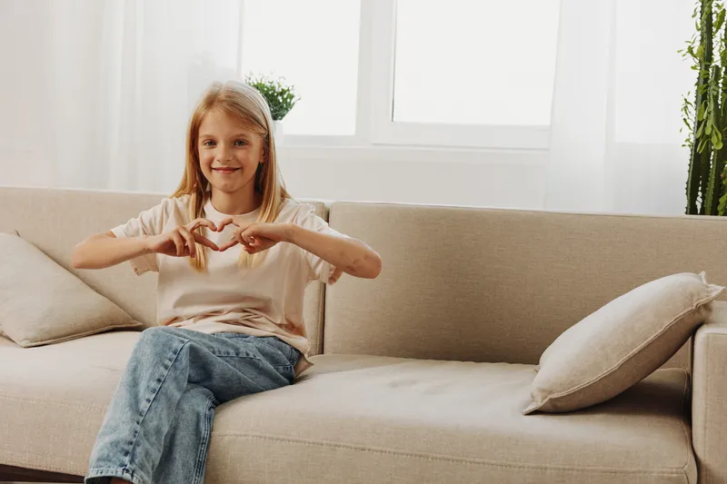 Smiling girl sitting on beige couch makes heart shape with hands in bright room. Concept of love, happiness, family, and positive emotions at home.