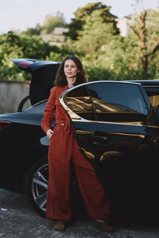 Woman in rust jumpsuit standing beside a sleek car outdoors, conveying modern lifestyle, confidence, and urban chic, with natural light, calm surroundings, and candid atmosphere