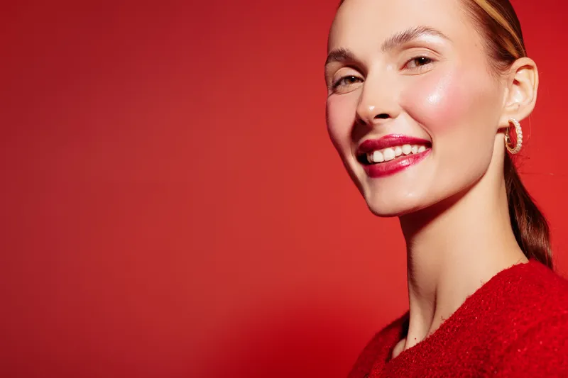 Smiling woman with red lipstick and earrings poses on red background. Close-up portrait of happy female model with shiny skin and pulled back hair. Concept of beauty and confidence.