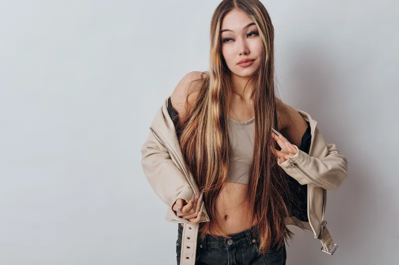 Young woman posing with long straight hair wearing casual jacket and crop top, looking sideways with confident expression on plain light background for fashion portrait.