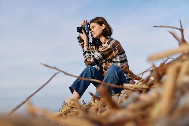 Photographer seated on driftwood wearing a plaid shirt, focused through a camera in an airy outdoor landscape by the beach, exploring light, color and composition in nature.