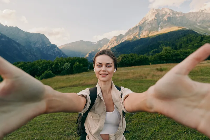 Young woman with brunette hair wearing a beige vest and white top takes a selfie with outstretched arms in a green meadow with mountains in the background.