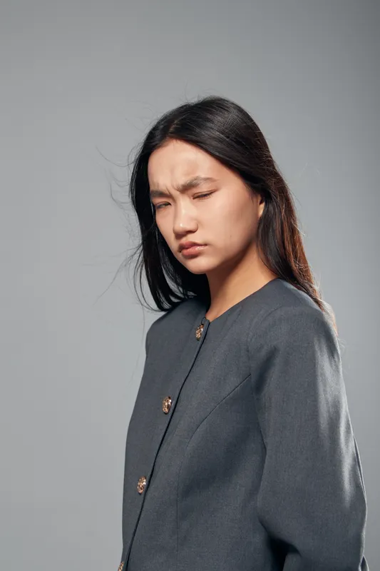 Young woman with long dark hair and gray blazer squinting eyes and frowning with displeased facial expression on gray background studio portrait.